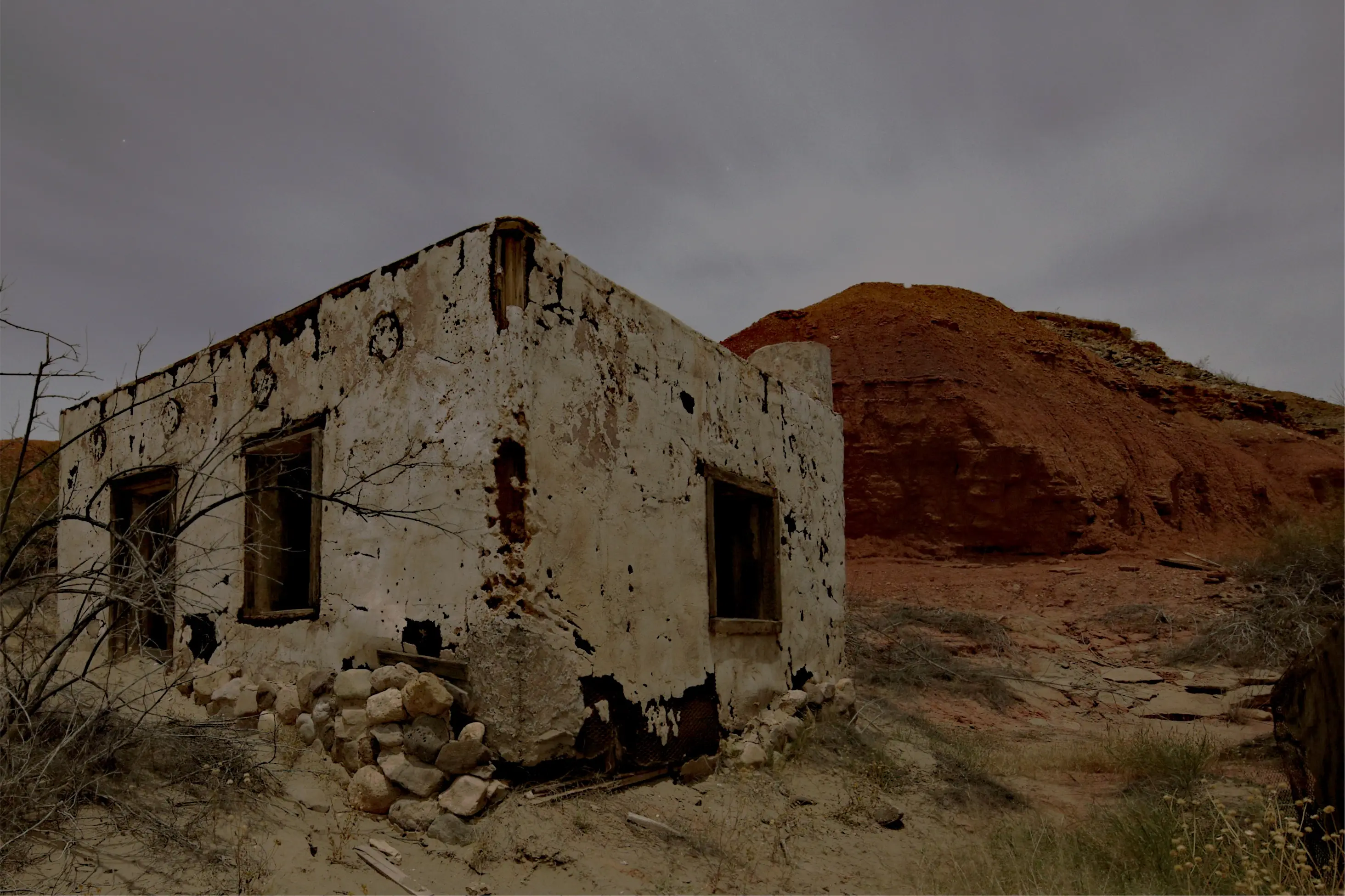 An abandoned adobe building in the Texas desert under a grey sky: whitewashed walls crumbling to brick, dark window openings, a pile of rocks at its base, sand and scrub around it, and a red clay hill behind. Selecting parts of it and moving the sliders beside it changes just those parts.