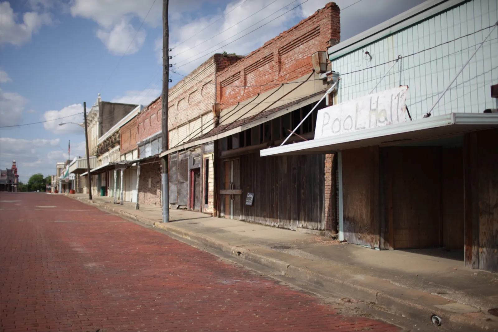 A small-town main street as shot: a row of old brick storefronts with awnings, one sign reading Pool Hall, under a blue sky with white clouds. Moving the sliders beside it changes how it looks.