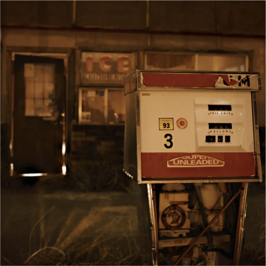 An old red and white gas pump at night, marked 3 and Super Unleaded, in front of a shuttered building with an ICE sign, warm-toned with a dark vignette.
